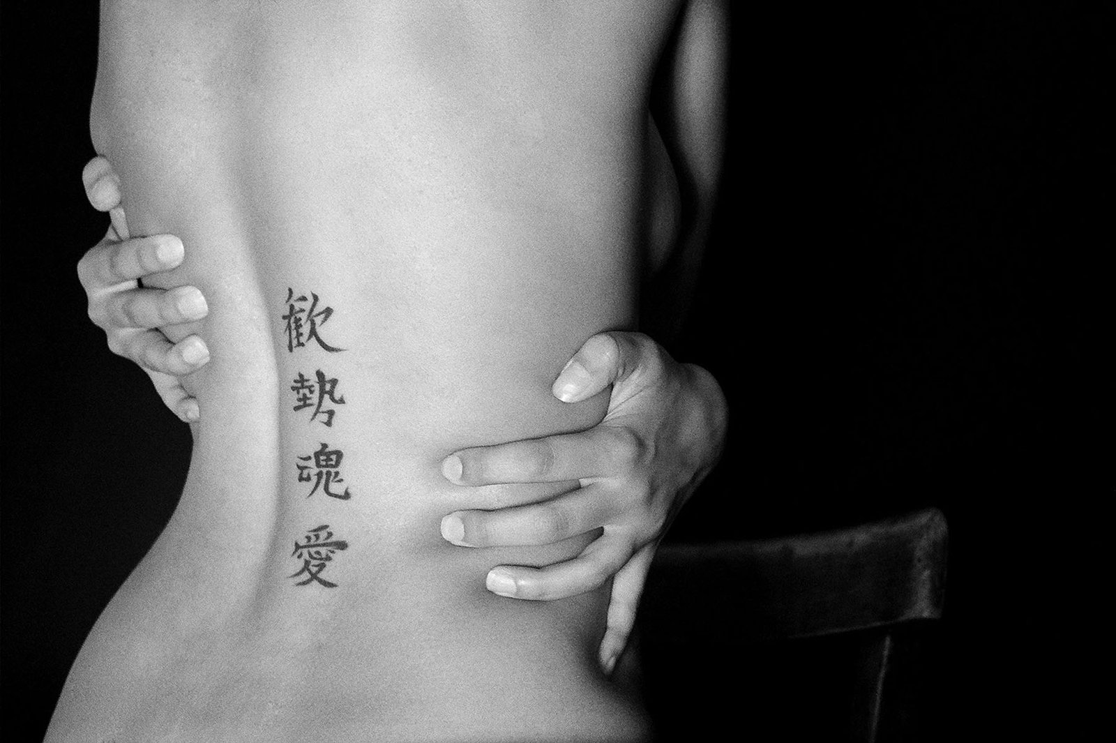 Black-and-white fine art portrait by Milan Stamenovic depicting a bare back held by two hands, marked with vertical calligraphic symbols, exploring intimacy, identity, and embodied resistance.