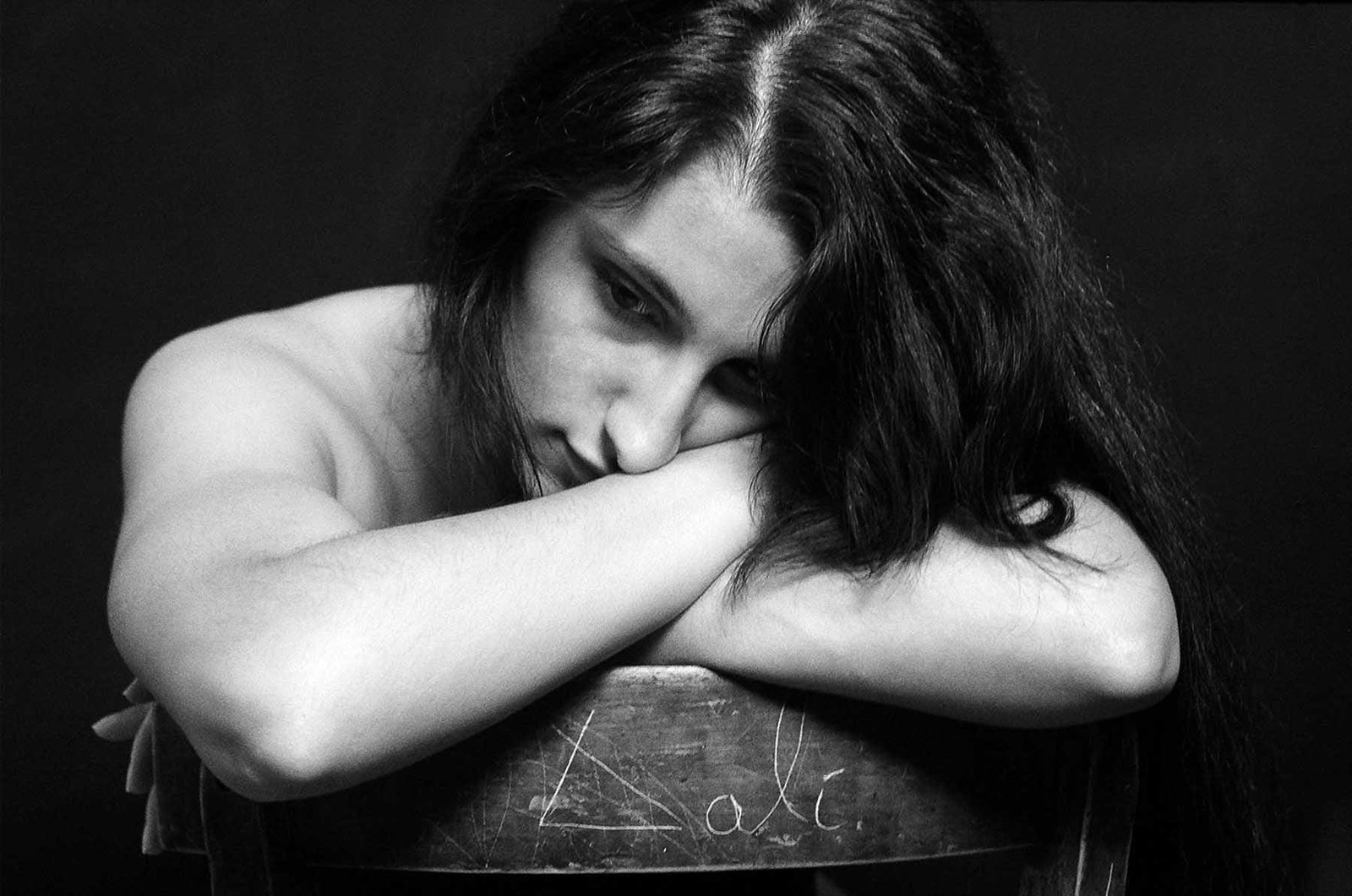Black-and-white fine art portrait by Milan Stamenovic showing a woman resting her head on folded arms against a wooden chair, expressing introspection, emotional tension, and quiet resistance.