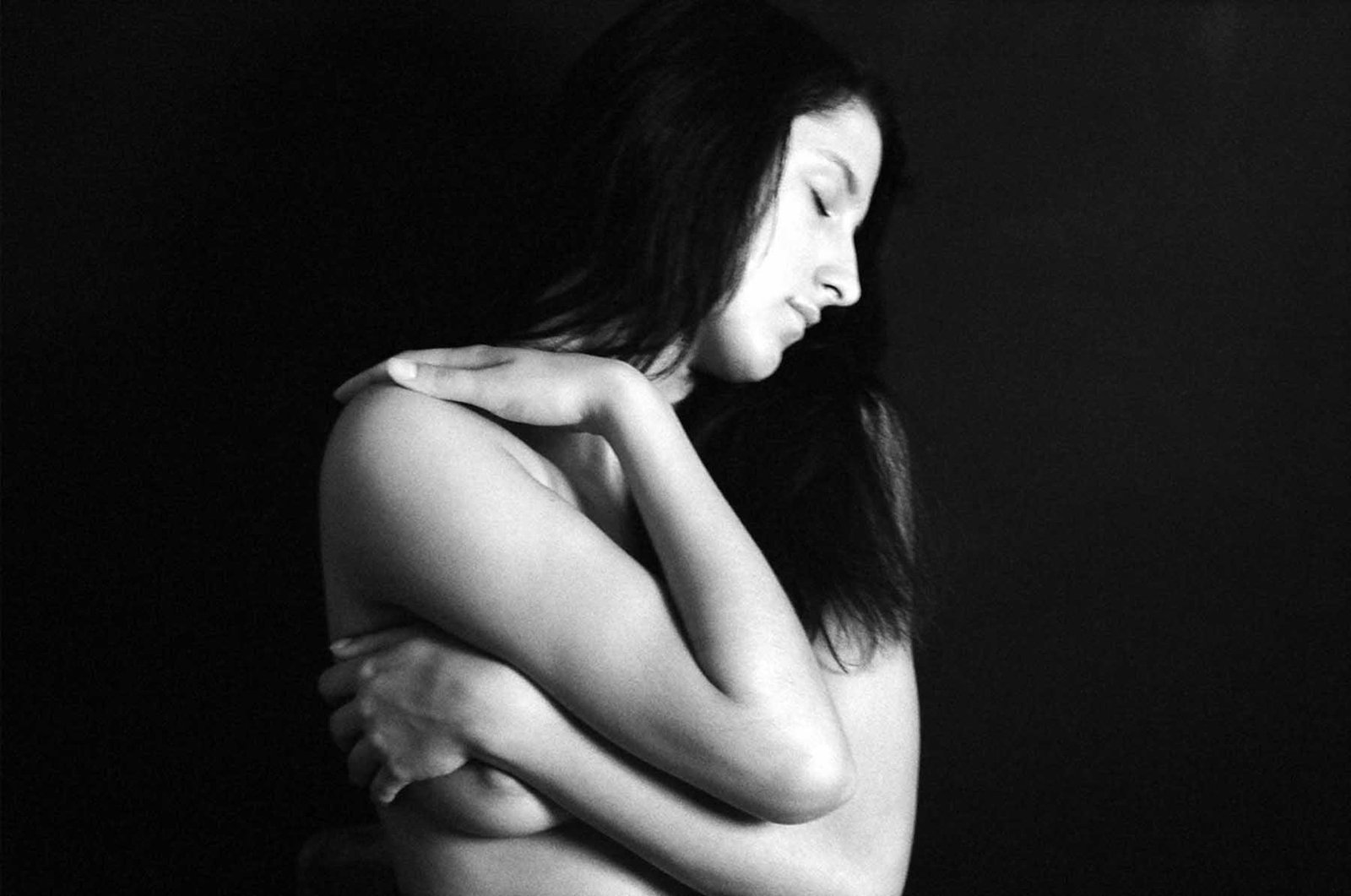 Black-and-white fine art portrait by Milan Stamenovic showing a woman embracing her own body with closed eyes, conveying intimacy, inner tension, vulnerability, and restrained emotional force.