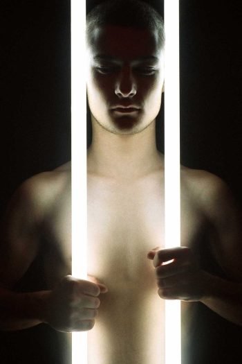Contemporary portrait photograph examining the human body held between illumination and collapse, where light becomes both revelation and destructive force.