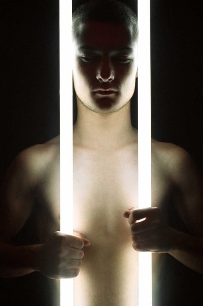 Contemporary portrait photograph examining the human body held between illumination and collapse, where light becomes both revelation and destructive force.