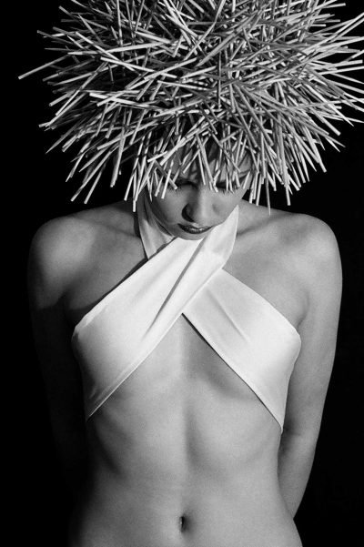 Black-and-white portrait photograph by Milan Stamenovic showing a figure crowned with a rigid, spiked structure, head lowered and body exposed against a dark background, evoking vulnerability, aftermath, and the residue of performance after the erosion of the authentic self (2004–2005).