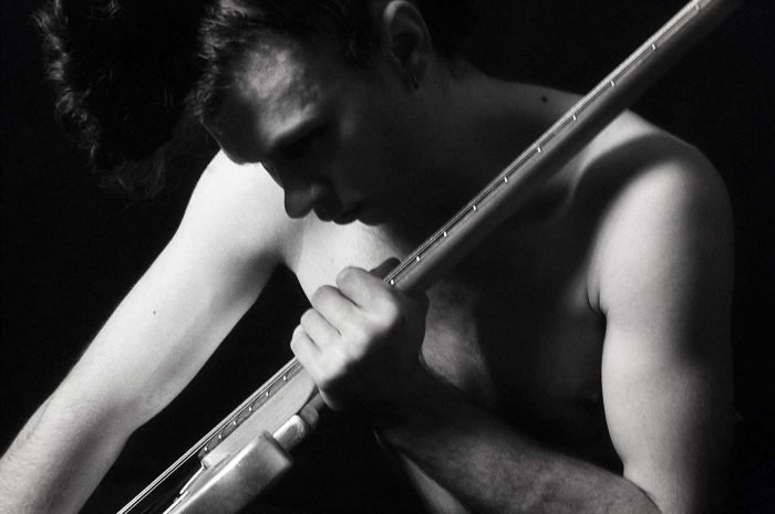 Black-and-white fine art portrait by Milan Stamenovic showing a nude male figure gripping the neck of a bass guitar, exploring cognitive tension, concentration, and the body as an instrument of thought and sound.