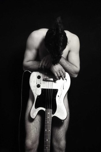 Black-and-white fine art portrait by Milan Stamenovic depicting a nude male figure leaning over an electric bass guitar, exploring physical burden, sonic weight, and the body as a vessel for sound.