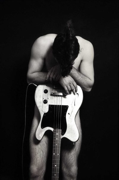 Black-and-white fine art portrait by Milan Stamenovic depicting a nude male figure leaning over an electric bass guitar, exploring physical burden, sonic weight, and the body as a vessel for sound.