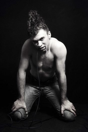 Black-and-white fine art portrait by Milan Stamenovic showing a kneeling male figure with a cable held in the mouth, exploring desire, electrical tension, and the body as a charged site of sonic intensity.