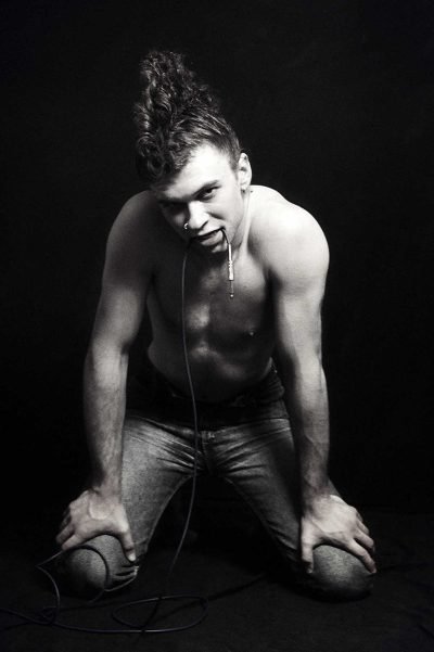 Black-and-white fine art portrait by Milan Stamenovic showing a kneeling male figure with a cable held in the mouth, exploring desire, electrical tension, and the body as a charged site of sonic intensity.