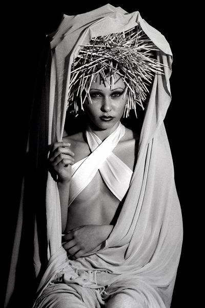Black-and-white portrait photograph by Milan Stamenovic depicting a figure draped in white fabric and crowned with a rigid, spiked headdress, examining the burden of imposed roles, scripted identity, and the erosion of authenticity within contemporary portrait photography (2004–2005).