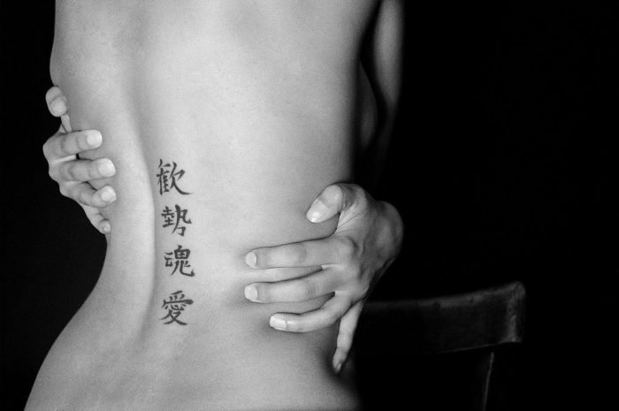 Black-and-white fine art portrait by Milan Stamenovic depicting a bare back held by two hands, marked with vertical calligraphic symbols, exploring intimacy, identity, and embodied resistance.