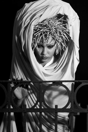 Black-and-white portrait photograph by Milan Stamenovic depicting a draped figure crowned with a rigid, spiked structure, leaning backward behind a metal railing, suggesting retreat from performed identity, hesitation, and the gradual erosion of the authentic self (2004–2005).