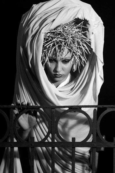 Black-and-white portrait photograph by Milan Stamenovic depicting a draped figure crowned with a rigid, spiked structure, leaning backward behind a metal railing, suggesting retreat from performed identity, hesitation, and the gradual erosion of the authentic self (2004–2005).