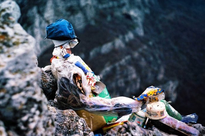 Fine art analog photograph depicting a human-like figure composed of discarded materials positioned within a rocky landscape, exploring human dominance, waste, and ecological consequence in a photographic work by Milan Stamenovic.