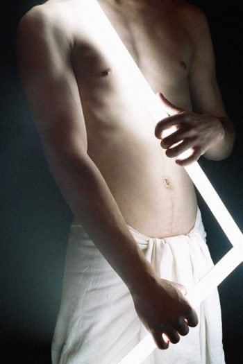 Contemporary portrait photograph examining light as an imposed condition, where illumination is held, crossed, and endured by the human body at the threshold of exposure.