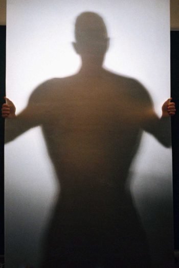 Portrait photograph of a blurred human silhouette gripping a translucent surface, examining containment, resistance, and embodied memory in Milan Stamenovic’s The Hidden Surface series.